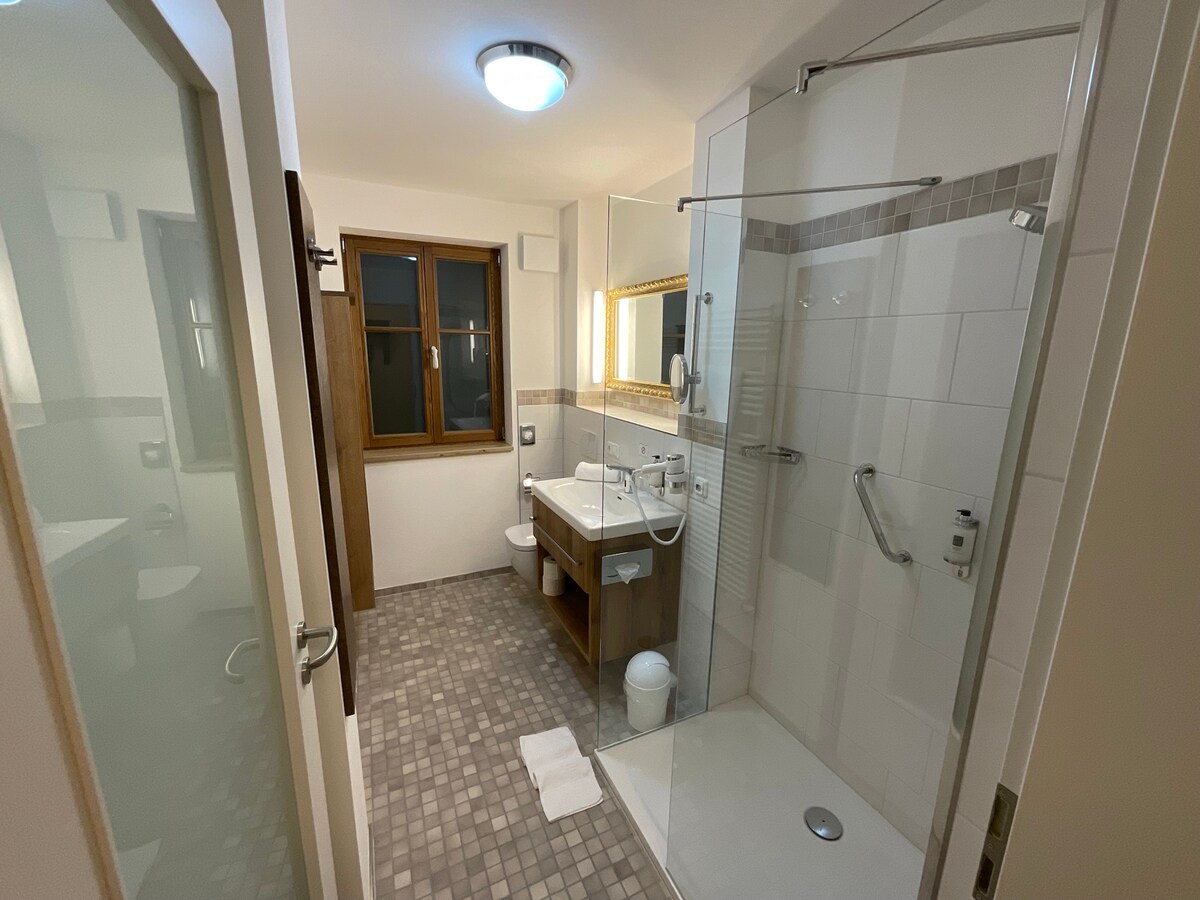 A modern bathroom features a walk-in shower with glass panels, a wooden vanity with a sink and mirrors. Natural light enters through a window, highlighting the neutral tile flooring. Essential fixtures are installed, offering a functional and clean space.