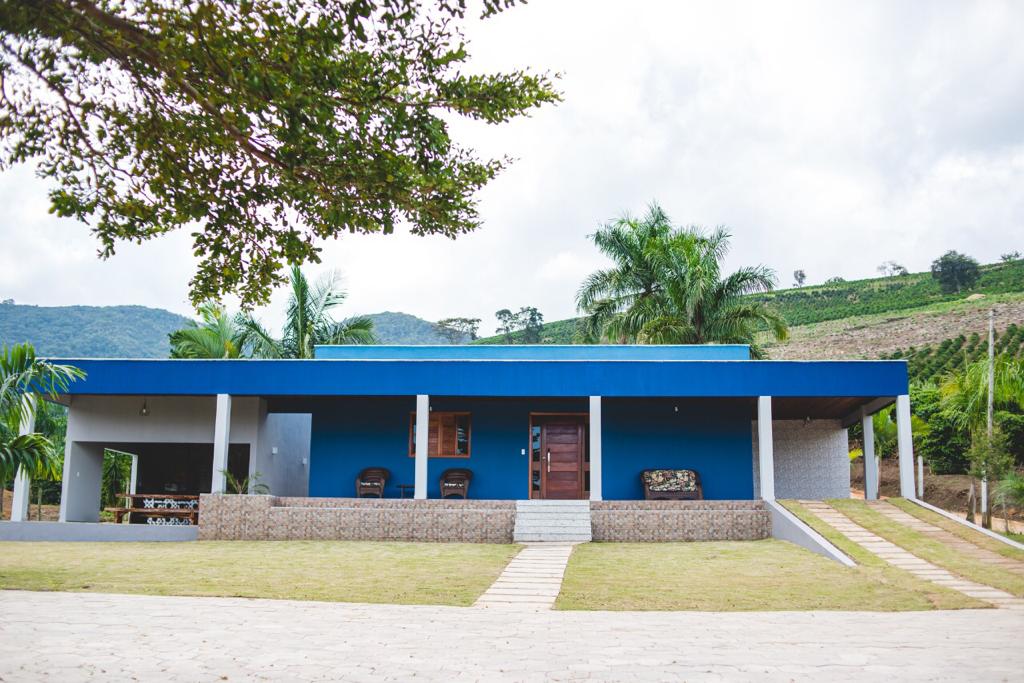 The exterior of the property features a modern, blue and gray façade complemented by lush greenery. A pathway leads to the entrance, while ample outdoor seating is visible under a shaded porch. The background showcases rolling hills, enhancing the serene rural setting.