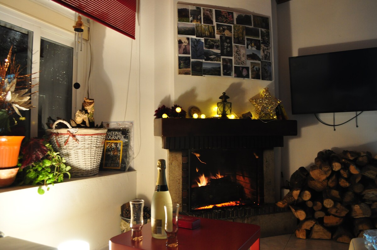 A welcoming living area is illuminated by warm candlelight from the fireplace. A collection of photographs covers the wall above the mantel, while a neatly stacked pile of firewood complements the cozy atmosphere. A red coffee table and sparkling glasses add a touch of charm.
