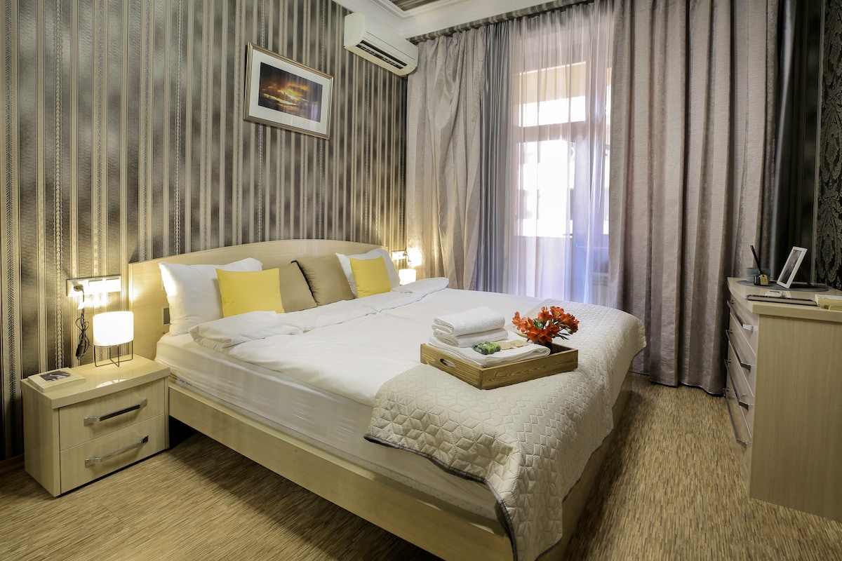 A well-lit bedroom features a spacious bed with white linens and decorative yellow pillows. Soft curtains frame the window, allowing natural light to filter in. A dresser and bedside tables complement the calming decor, while a touch of color is added by flowers on the tray.