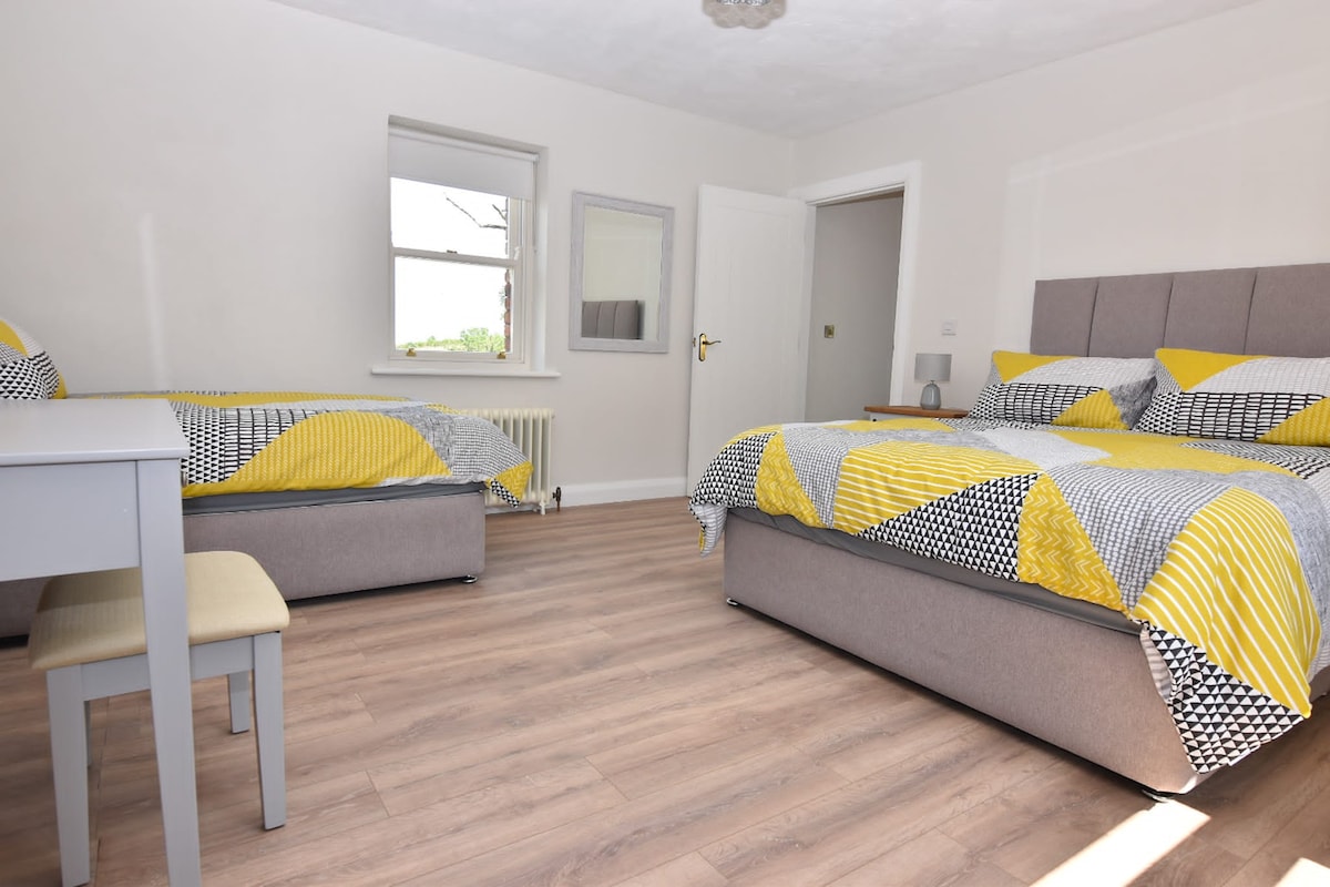 A well-lit bedroom features two beds dressed in patterned yellow and black linens. A desk and chair are positioned in one corner, while a window allows natural light to fill the space. Light wood flooring complements the neutral wall color, creating a fresh and inviting atmosphere.