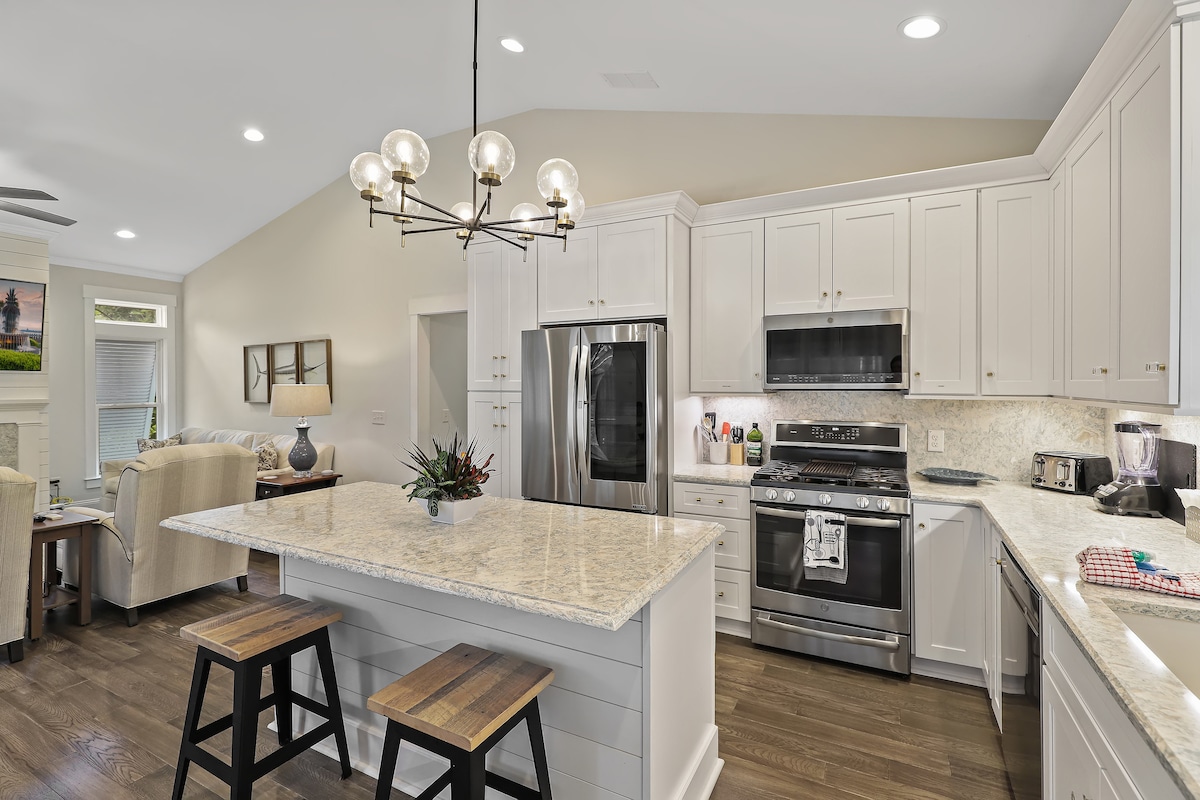 The kitchen features light-colored cabinetry and quartz countertops, complemented by stainless steel appliances. A large island with seating for two is available. Soft ambient lighting from a modern chandelier enhances the space, creating a warm atmosphere that connects to the adjacent living area.