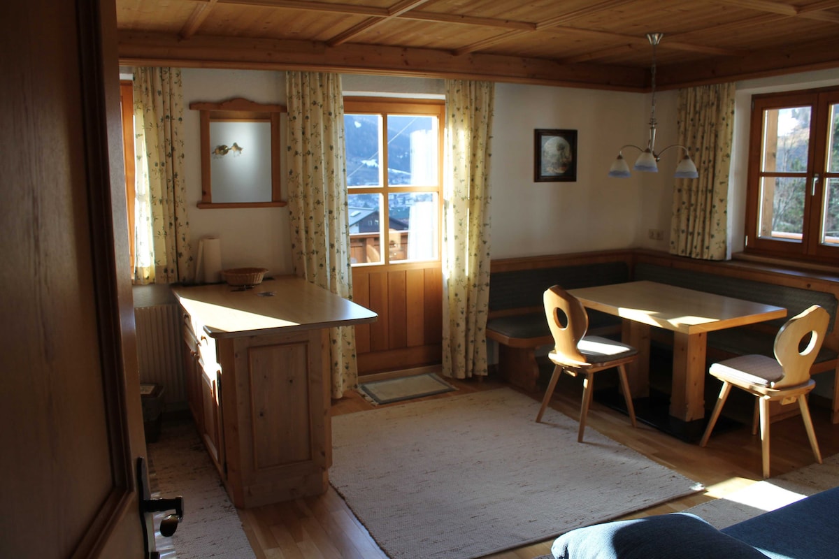 A dining area is illuminated by natural light flowing through large windows. A wooden table with four chairs sits beside a cozy bench. The room features a warm wood ceiling, light-colored walls adorned with patterned curtains, and a rug enhancing the living space.