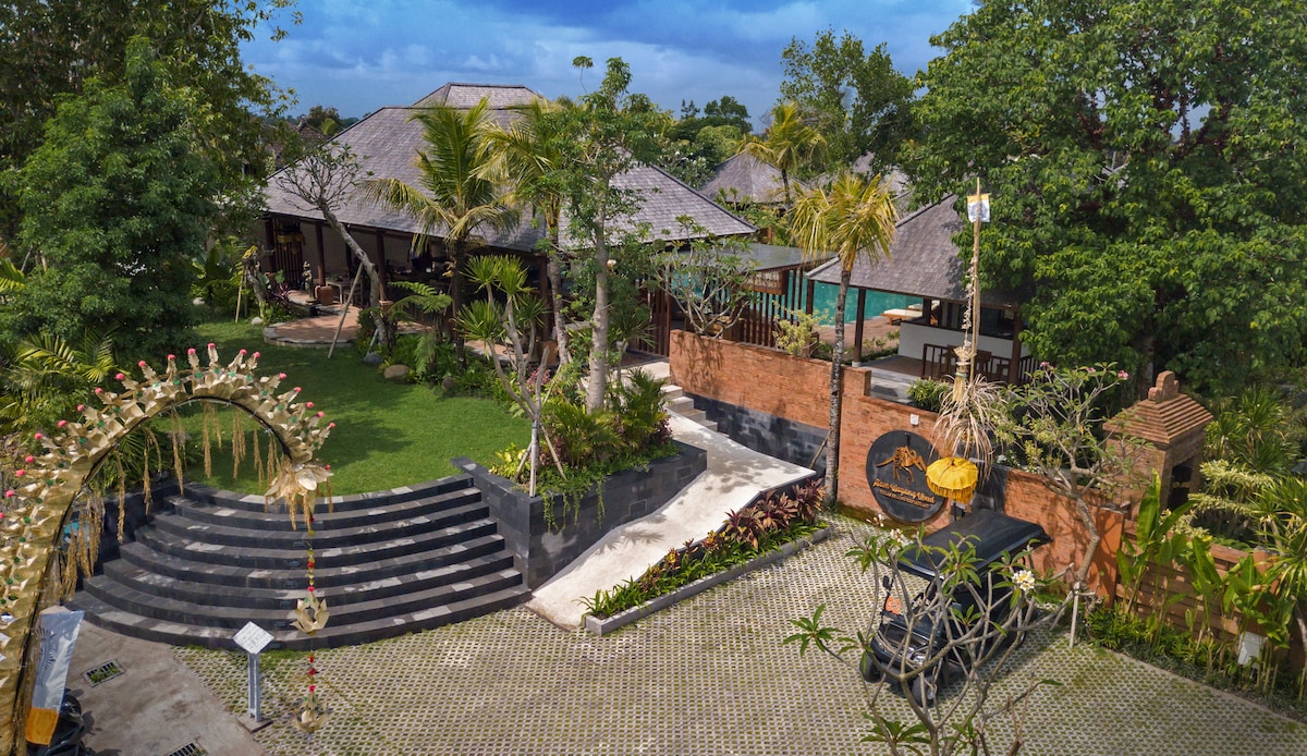 A spacious outdoor area is depicted, featuring lush green landscaping and various palm trees. A large pool is visible in the background, alongside traditional Balinese architecture with thatched roofs. Stone pathways and decorative elements enhance the tranquil atmosphere of the property.