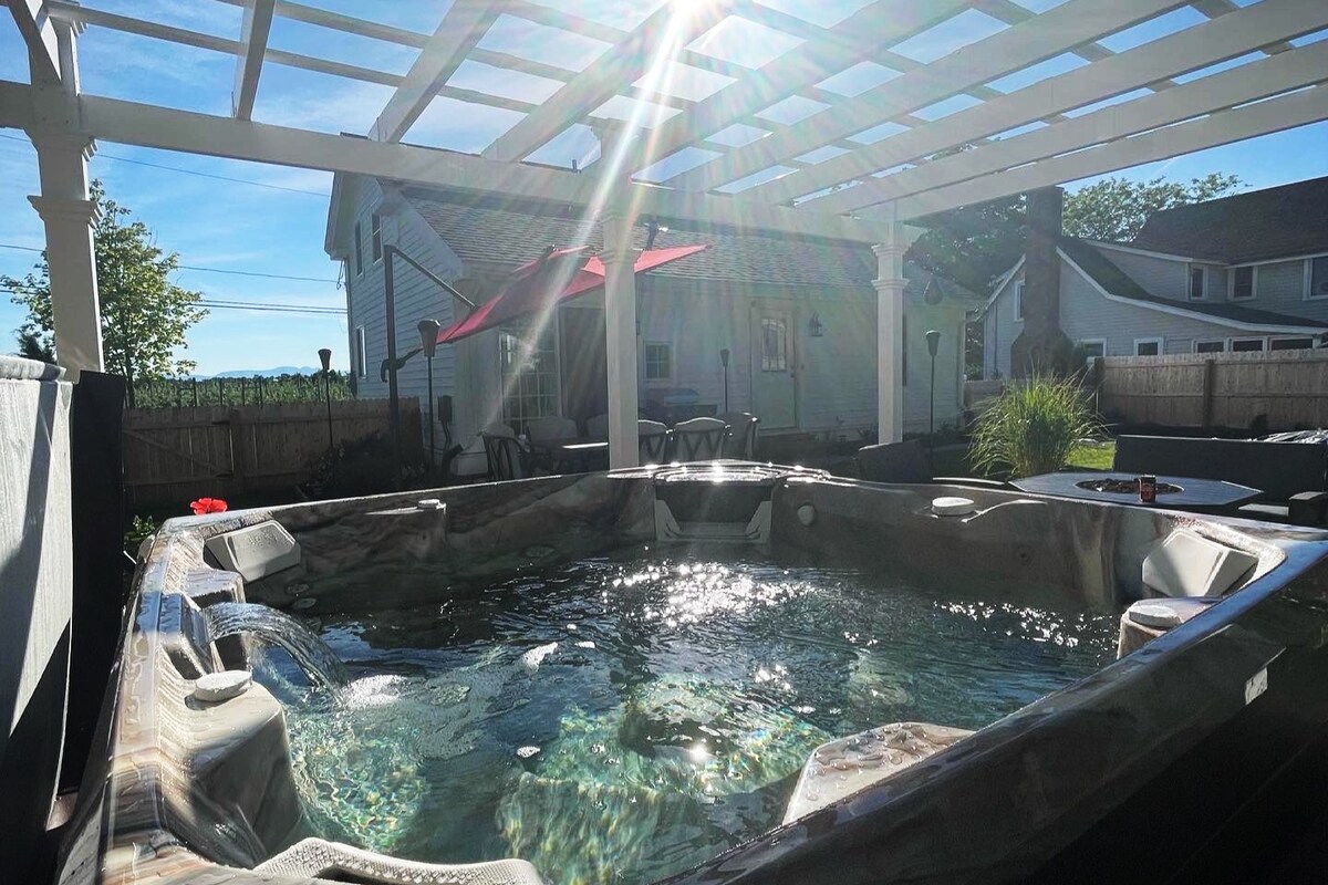 A spacious hot tub is situated beneath a pergola, surrounded by flowing water features. Sunlight filters through the structure, creating a warm ambiance. Dining areas with an umbrella can be seen in the background, along with lush greenery and a fence securing the private outdoor space.