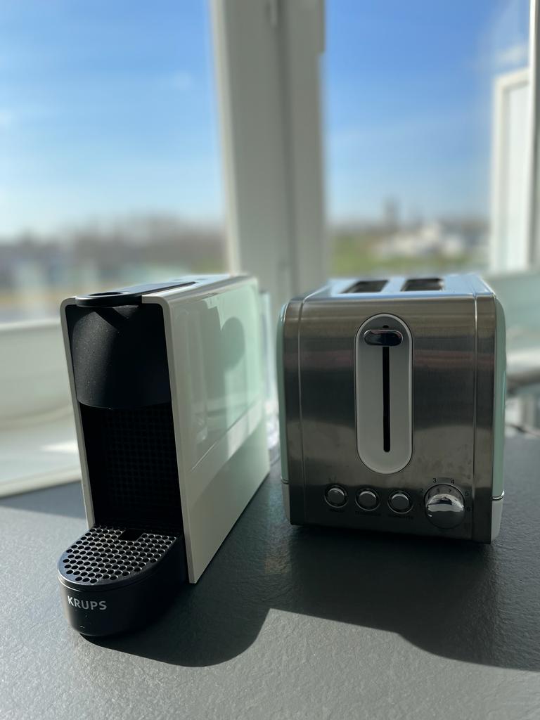 A coffee machine and a toaster are placed on a countertop. The coffee machine features a sleek white design, while the toaster has a brushed metal finish. Natural light streams through a nearby window, illuminating the countertop and appliances.