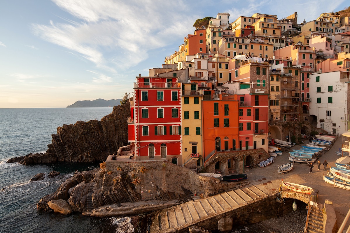 The vibrant colors of Riomaggiore's buildings contrast against the clear blue sea. The structure's facades display shades of orange, red, yellow, and green, while fishing boats rest along the rocky shore. The scene captures the charm of this coastal village at sunset.