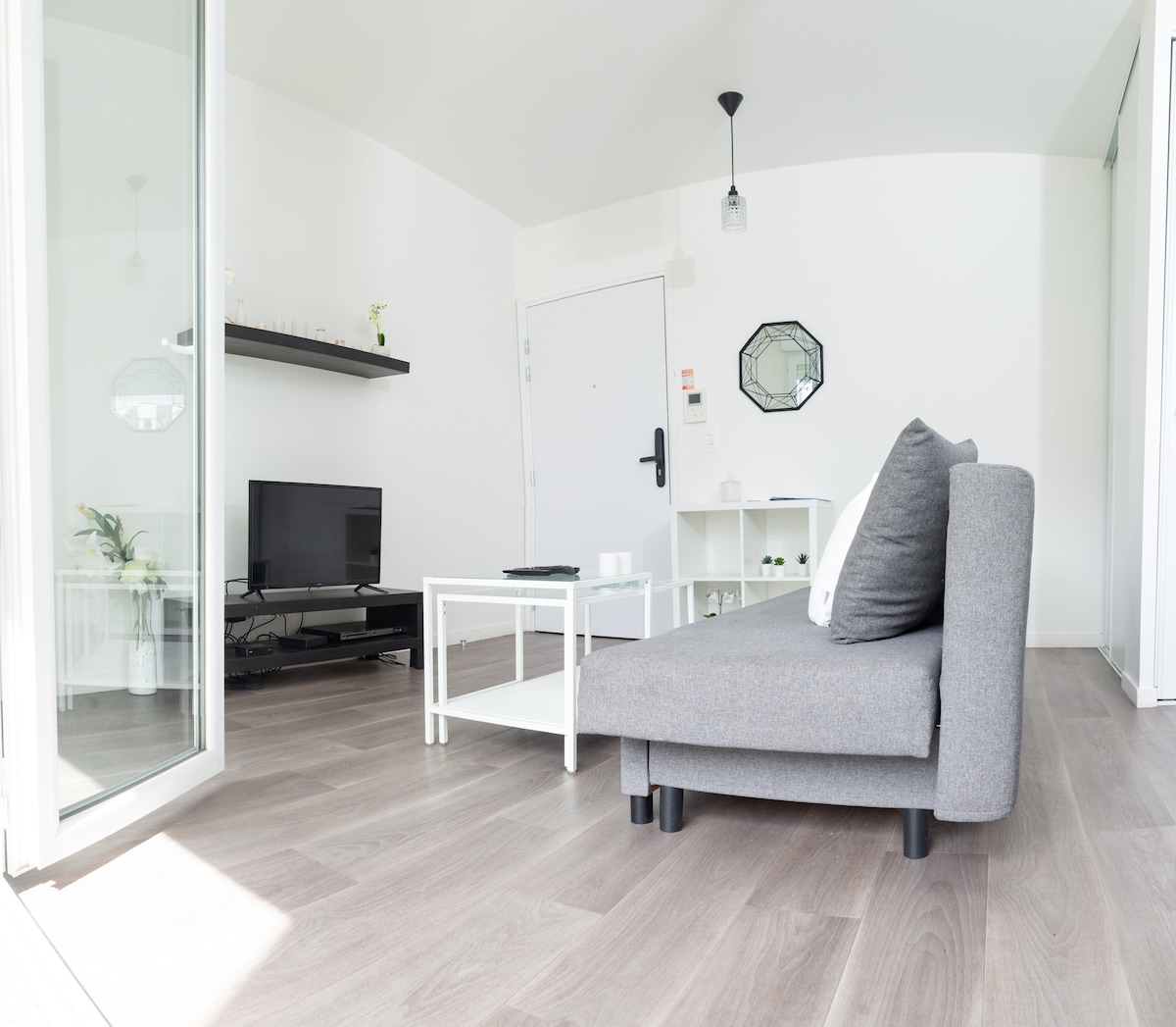 A spacious living area features a grey sofa positioned toward a wall-mounted television on a sleek shelf. A white coffee table sits in front of the sofa, complemented by minimal decorative items. Natural light enters through large windows, enhancing the bright and open layout.