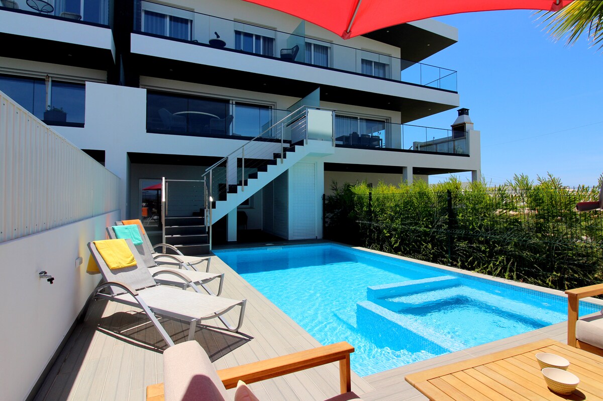 A modern villa features a private swimming pool framed by sun loungers and a wooden deck. An outdoor seating area is visible beside the pool, and a staircase leads to the upper levels. Greenery provides a natural backdrop, enhancing the outdoor space.