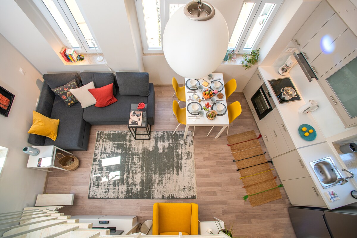 An aerial view of a bright, open living space features a cozy gray sofa adorned with colorful cushions. A dining table is set for a meal with yellow chairs, and a stylish rug anchors the area. Light streams in through multiple windows, enhancing the modern kitchen's atmosphere.