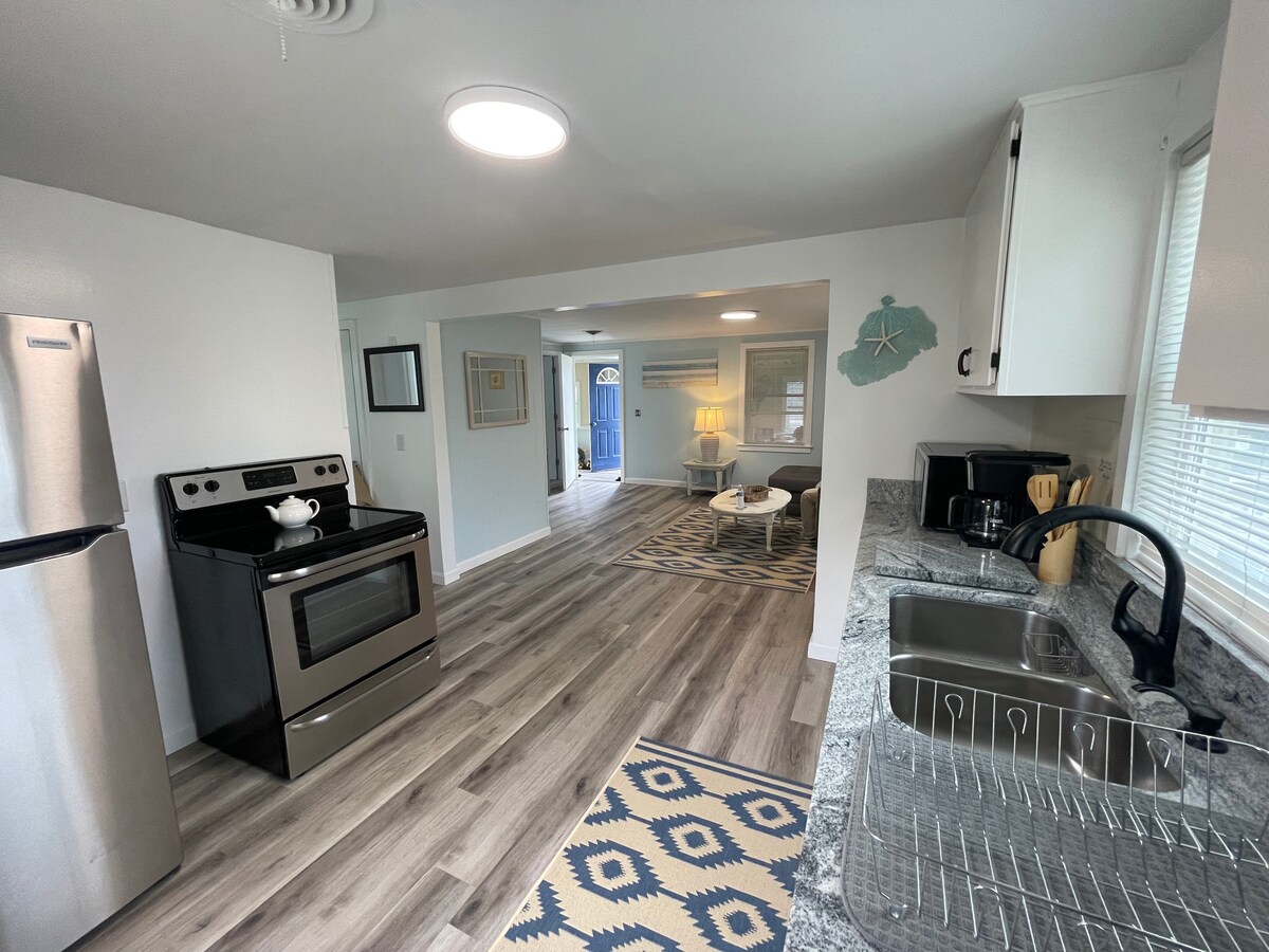 A modern kitchen area features stainless steel appliances, including a stove and refrigerator. Gray countertops and a patterned area rug contribute to a clean and functional space. Natural light fills the room, highlighting the open layout that connects to a living area with a blue door visible in the background.