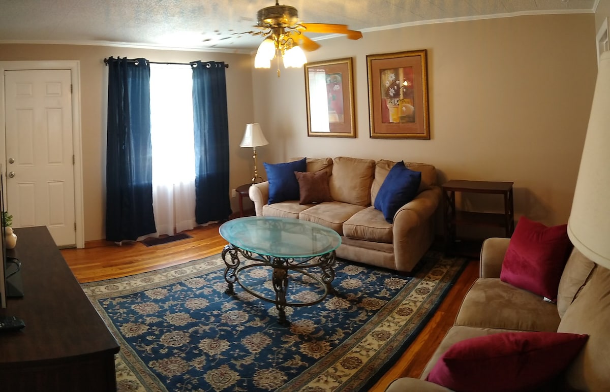 A comfortable living area is shown with a large sectional sofa adorned with blue and red cushions. A round glass coffee table sits atop a patterned area rug, while framed artwork decorates the walls. Natural light enters through the window, complemented by dark curtains.