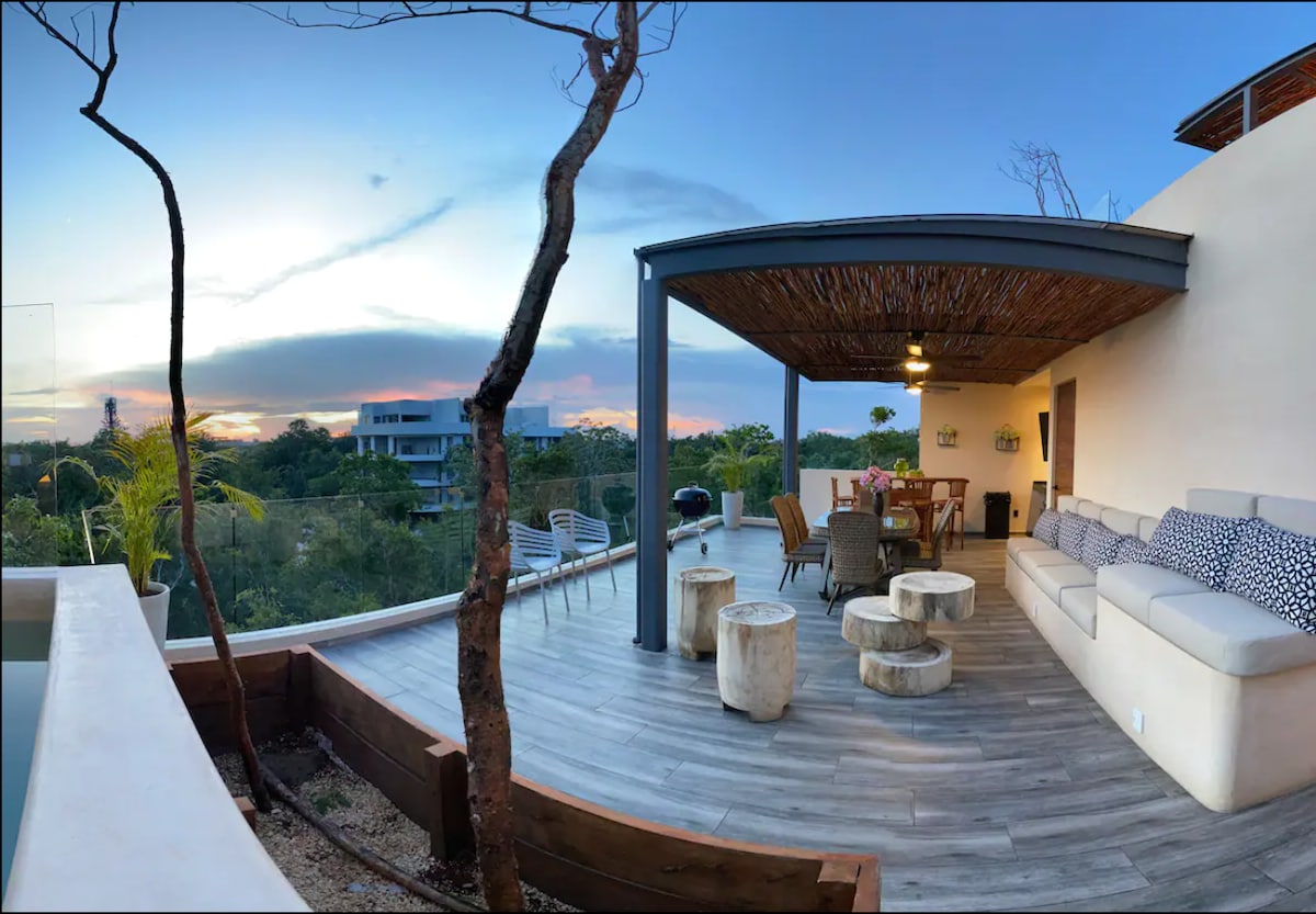 An outdoor terrace features a large, shaded seating area with a spacious couch and rustic wooden stools. A dining table is placed nearby, surrounded by lush greenery and a view of the tranquil surroundings, highlighting the connection with nature as the sun sets in the background.