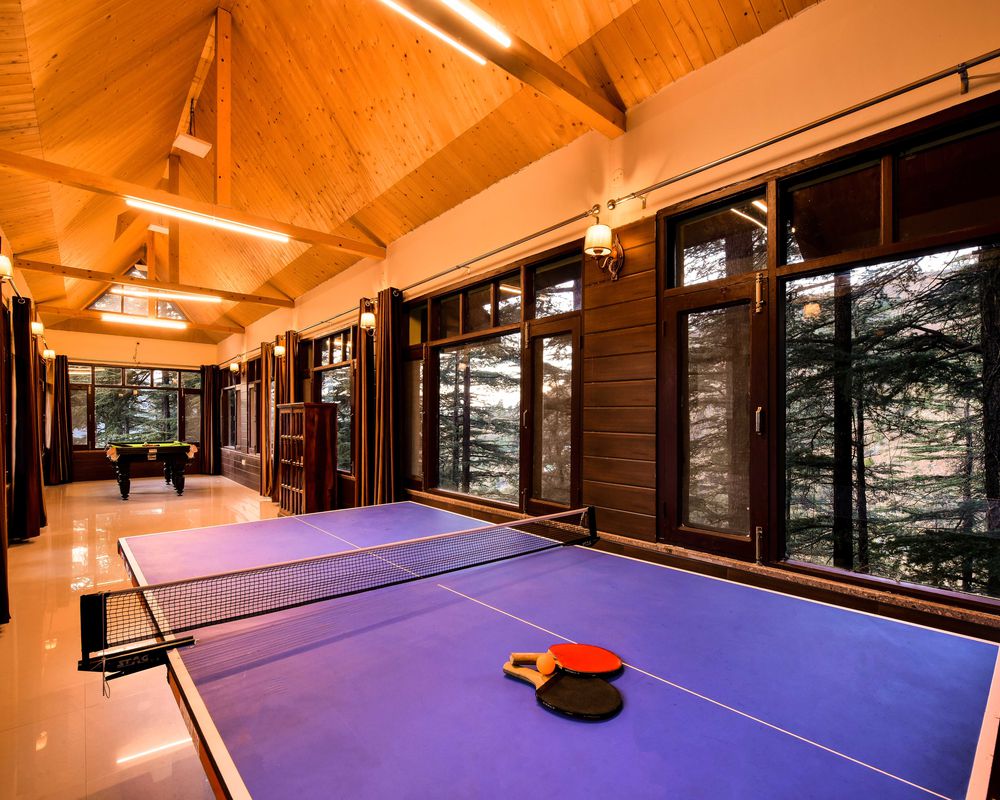A spacious games area features a blue table tennis table with paddles and a ball positioned on it. Large windows allow natural light to stream in, providing views of surrounding trees. The wooden ceiling adds warmth to the bright interior.
