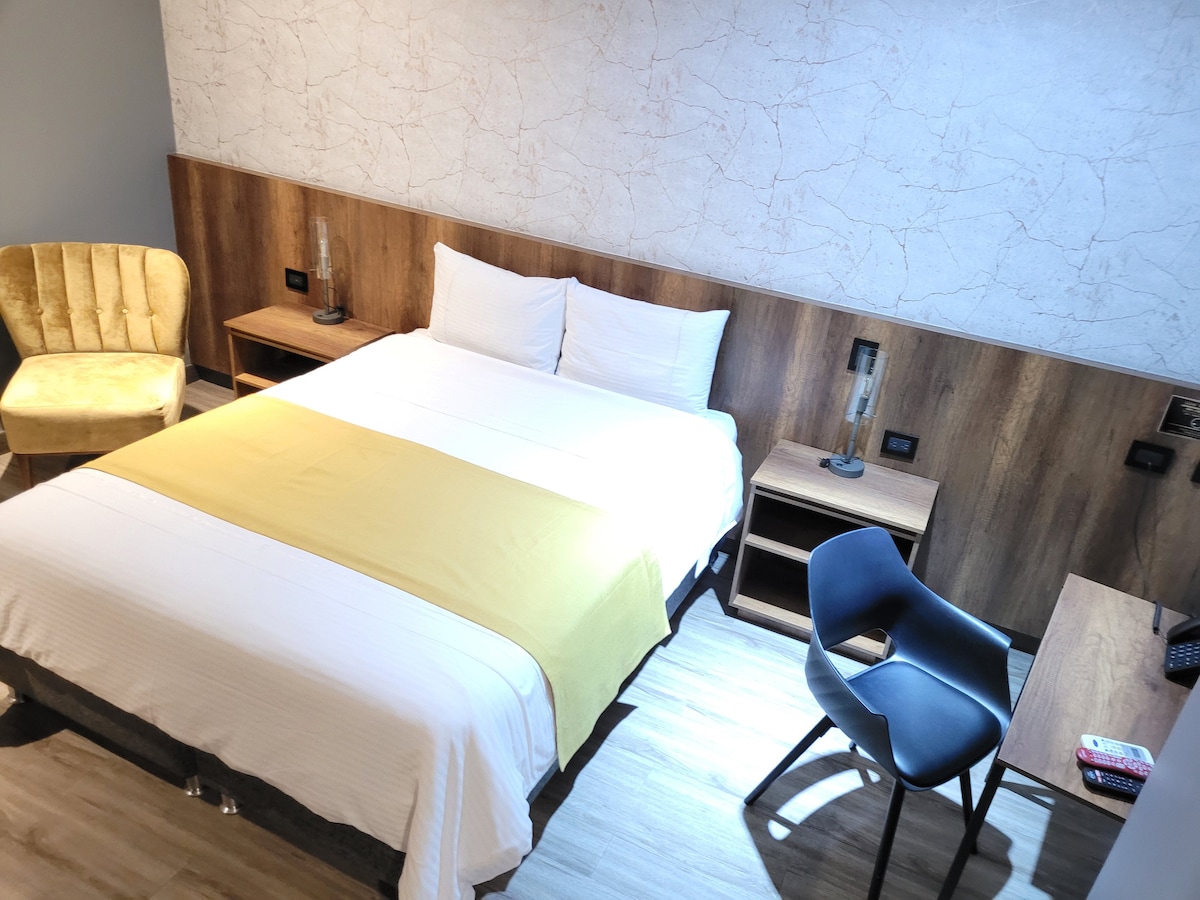 A spacious guest room features a large bed with a crisp white cover and a yellow accent. Wooden elements provide a warm touch, while a comfortable chair and desk are positioned nearby. Natural light illuminates the space, and bedside lamps add a contemporary vibe.