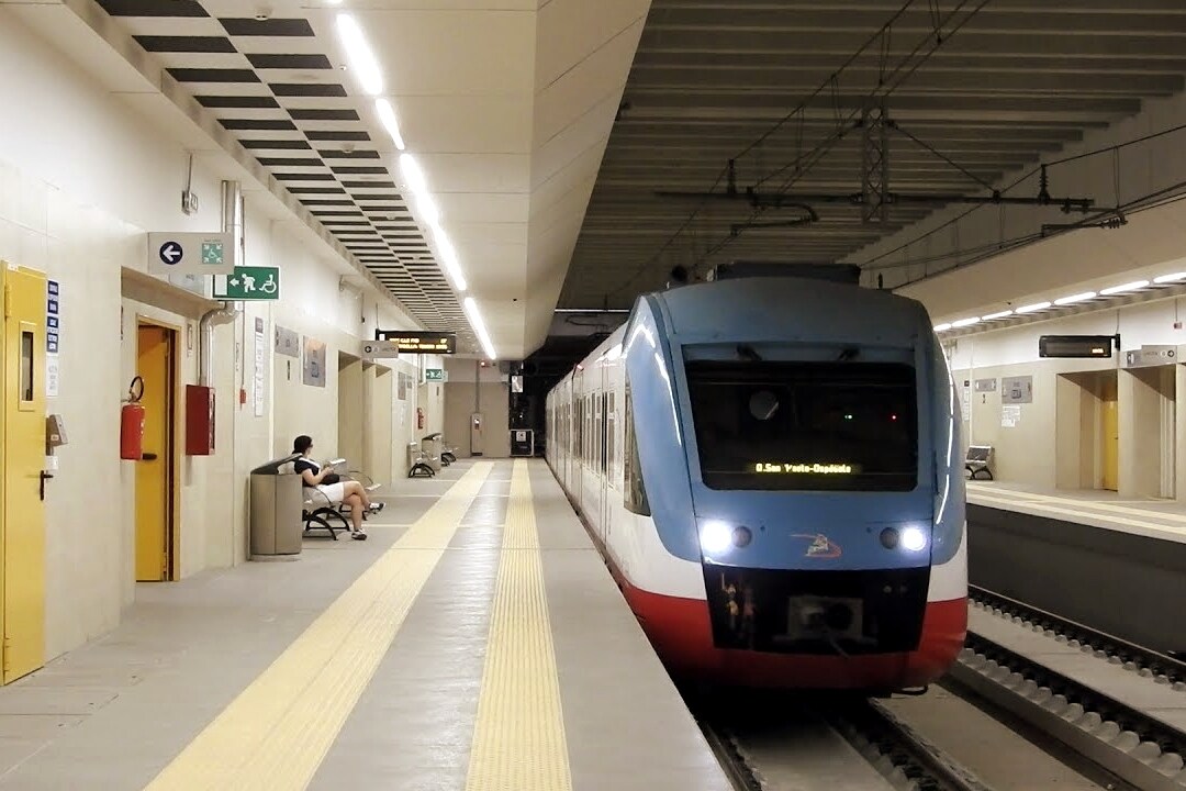 A train platform is depicted, featuring a train in motion on the left side. Yellow tactile paving guides individuals along the platform. Seating is available along the wall, with a passenger seated in the distance. Station signage indicates directions and amenities.