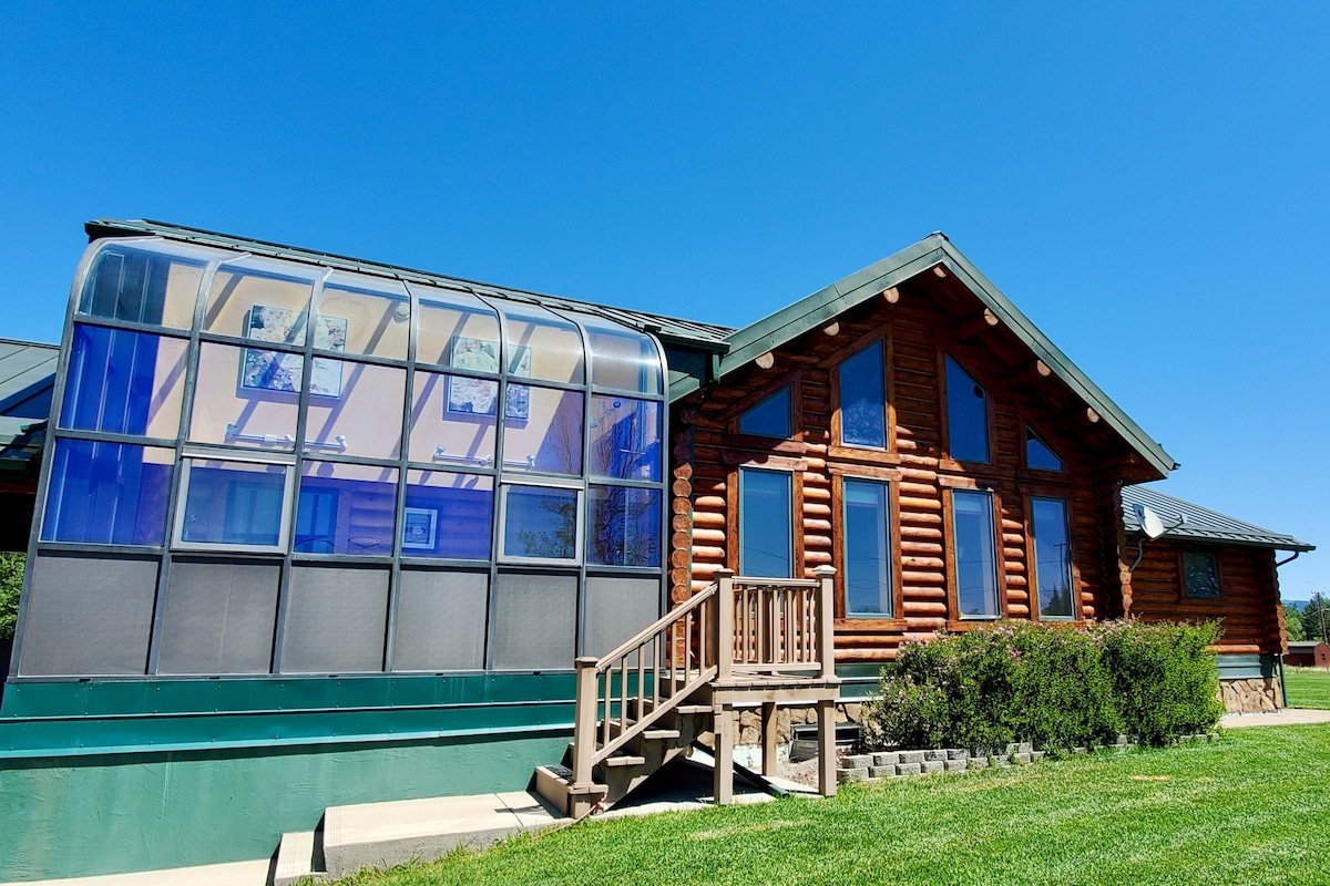The exterior of a three-level log home is showcased with large, angled windows and a sunroom extension. The inviting wooden entrance steps lead up to a green-painted porch. Surrounding the home is a well-maintained lawn and clear blue sky, enhancing the tranquil outdoor setting.