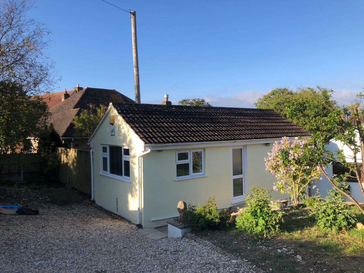 The exterior of a yellow studio apartment is shown, featuring a sloped roof and multiple windows. A gravel pathway leads to the entrance, surrounded by greenery. Sunlight highlights the fresh facade, and a power line is visible above the structure.