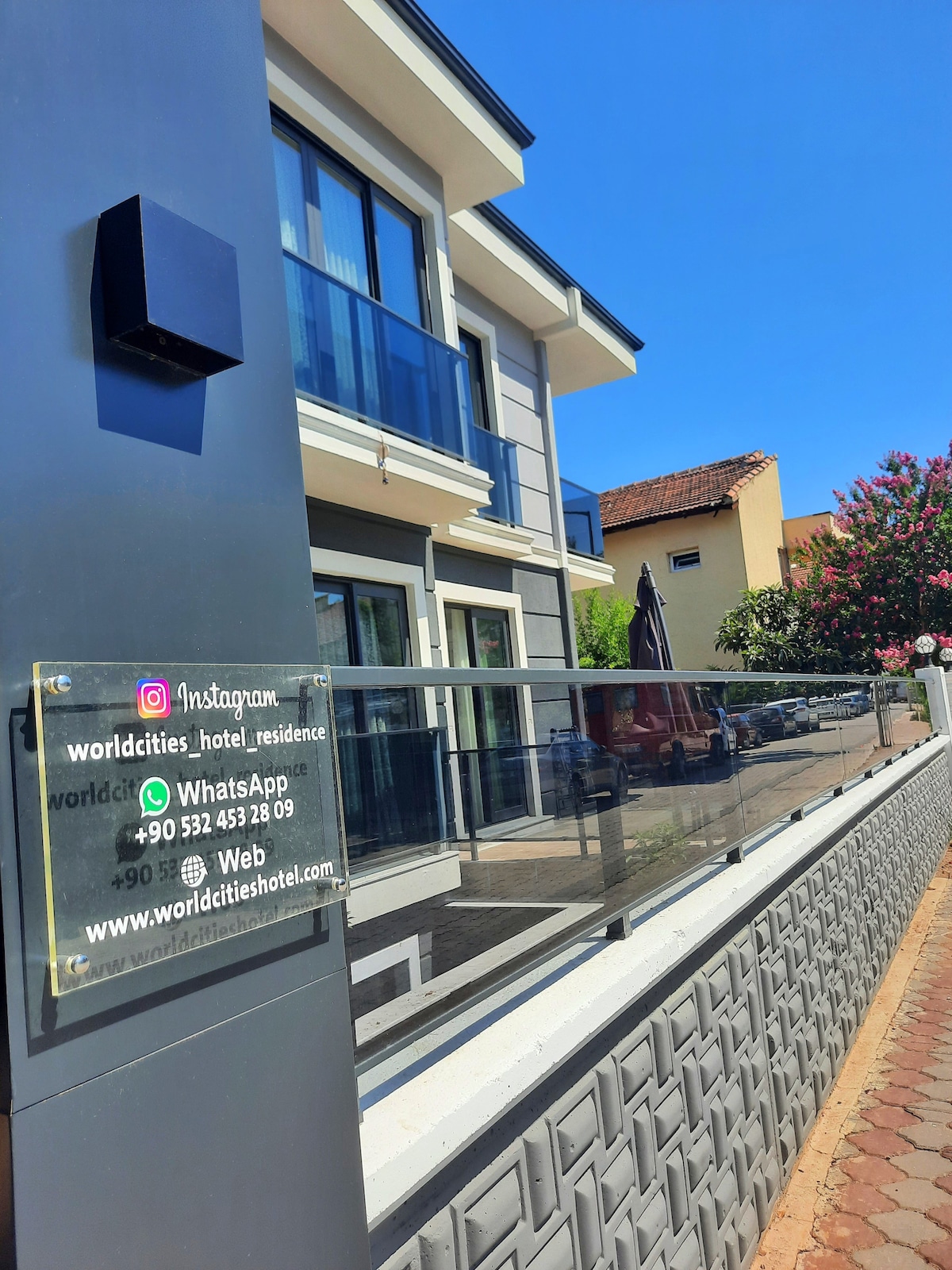 The exterior of a modern apartment hotel is depicted, featuring multiple large windows and a spacious balcony. A glass railing is visible in the foreground, enhancing the sleek design. The building's contemporary architecture is framed by a clear blue sky and lush greenery.