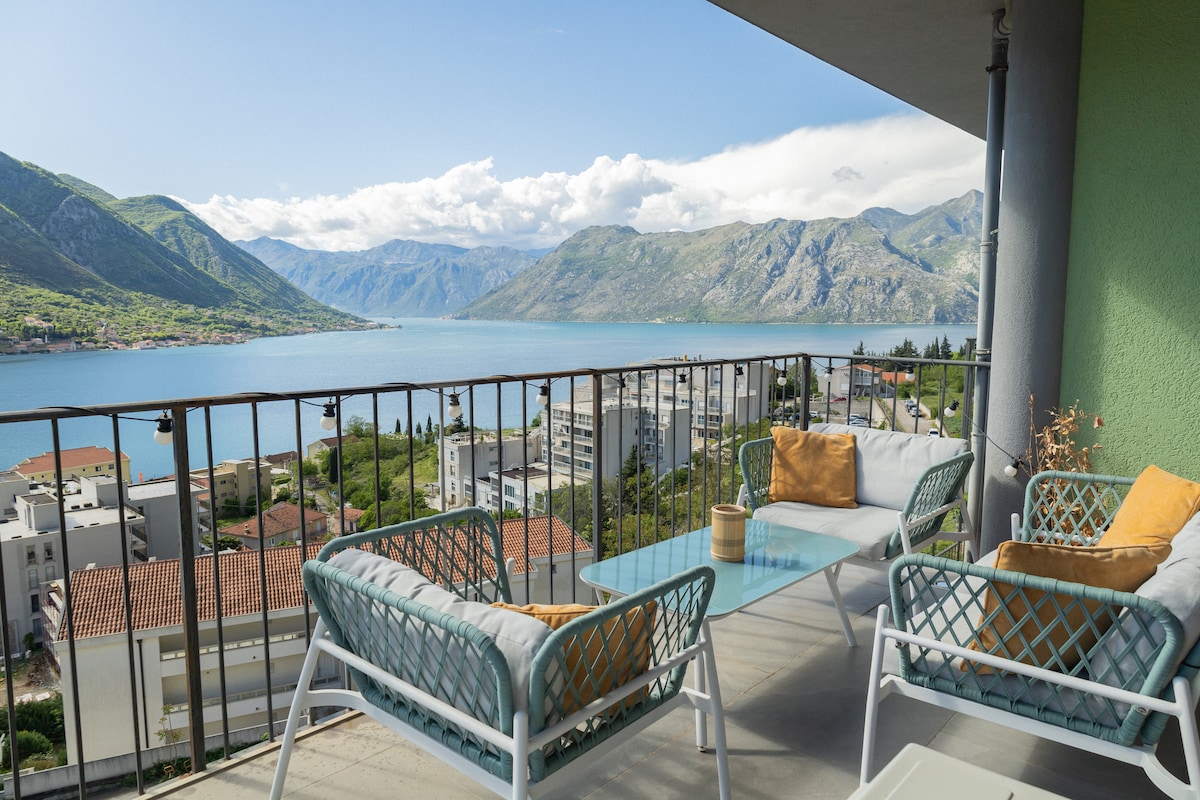 A spacious balcony is furnished with two cozy armchairs and a glass-topped table, complemented by decorative cushions. The picturesque view of Kotor Bay and surrounding mountains is prominently featured, with a clear sky adding to the serene atmosphere.