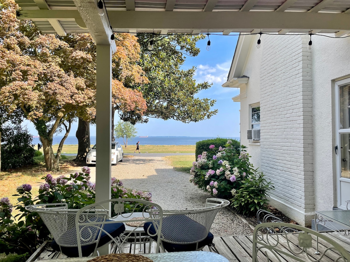 A covered porch overlooks a tranquil landscape, featuring lush greenery and blooming hydrangeas. The bay is visible in the distance, framed by trees with autumn foliage. Outdoor seating is arranged under the pergola, inviting relaxation while enjoying the scenic view.