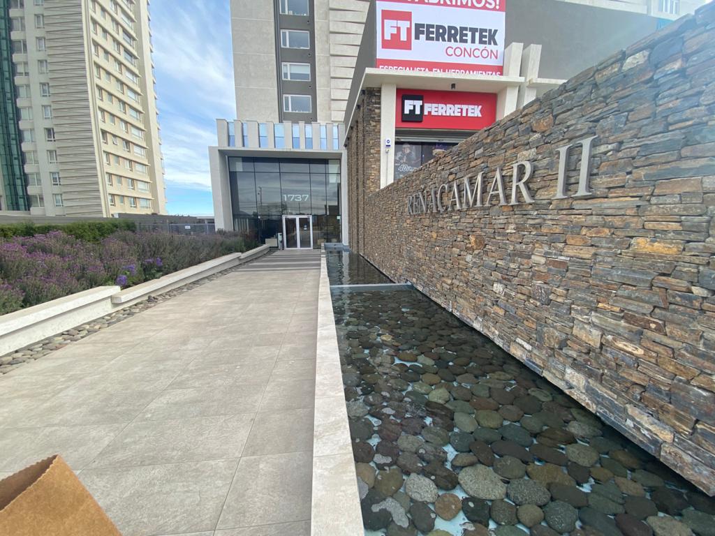 The building entrance is framed by a contemporary stone wall, with water quietly reflecting the surroundings. A clean walkway leads to the glass doors, with vibrant plants visible on either side, providing a welcoming approach to the loft.