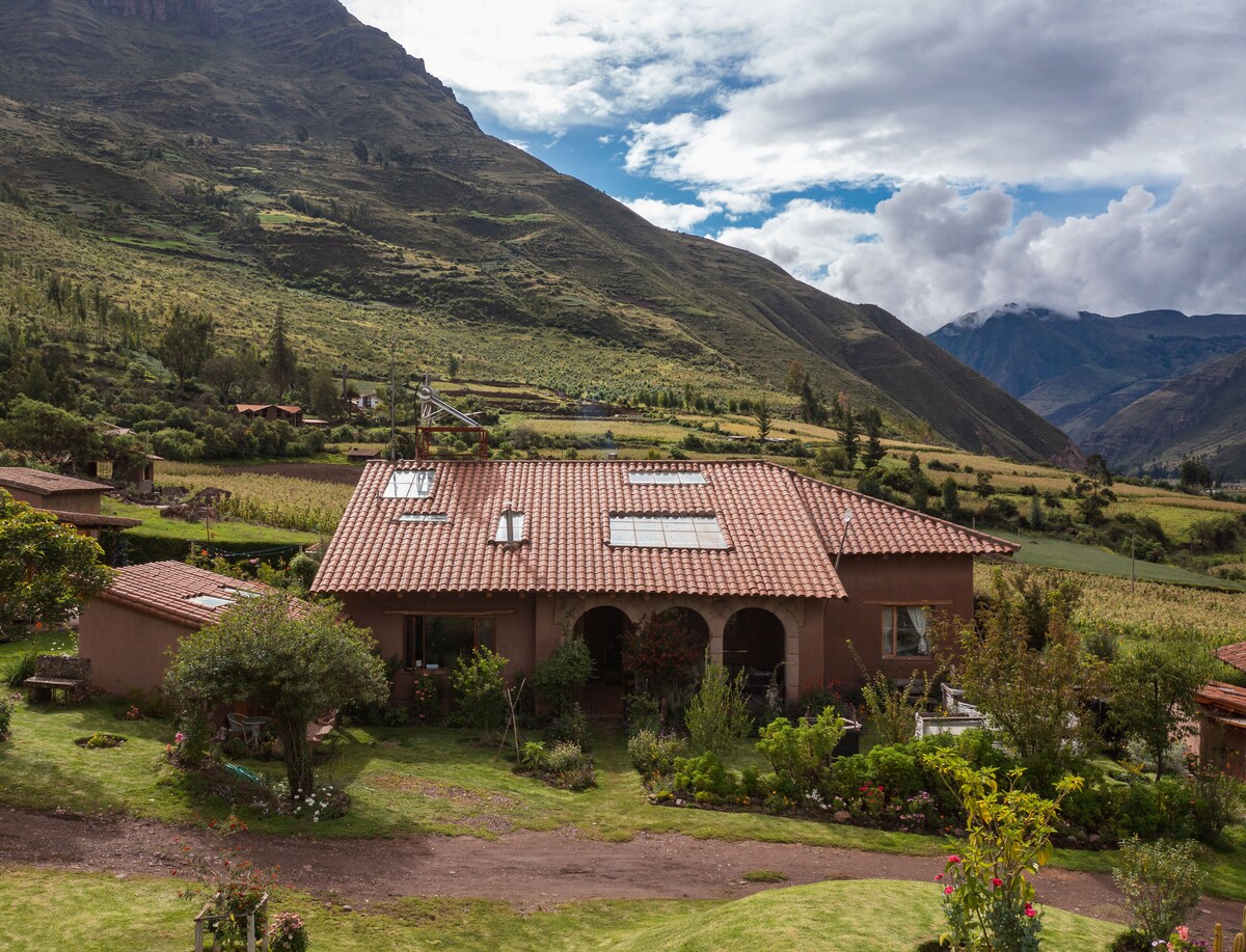 The cabin features a rustic exterior with a tiled roof, surrounded by well-maintained gardens. Large windows and sky lights offer natural light, while the backdrop of vibrant hills creates a serene atmosphere. A pathway leads to the entrance, enhancing the welcoming nature of the property.