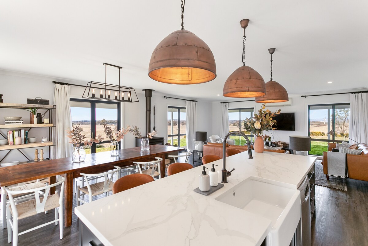 A spacious dining and kitchen area features a large marble island with a modern sink. Pendant lights with copper shades illuminate the space, complemented by a wooden dining table and white chairs. Large glass doors provide abundant natural light and views of the surrounding landscape.