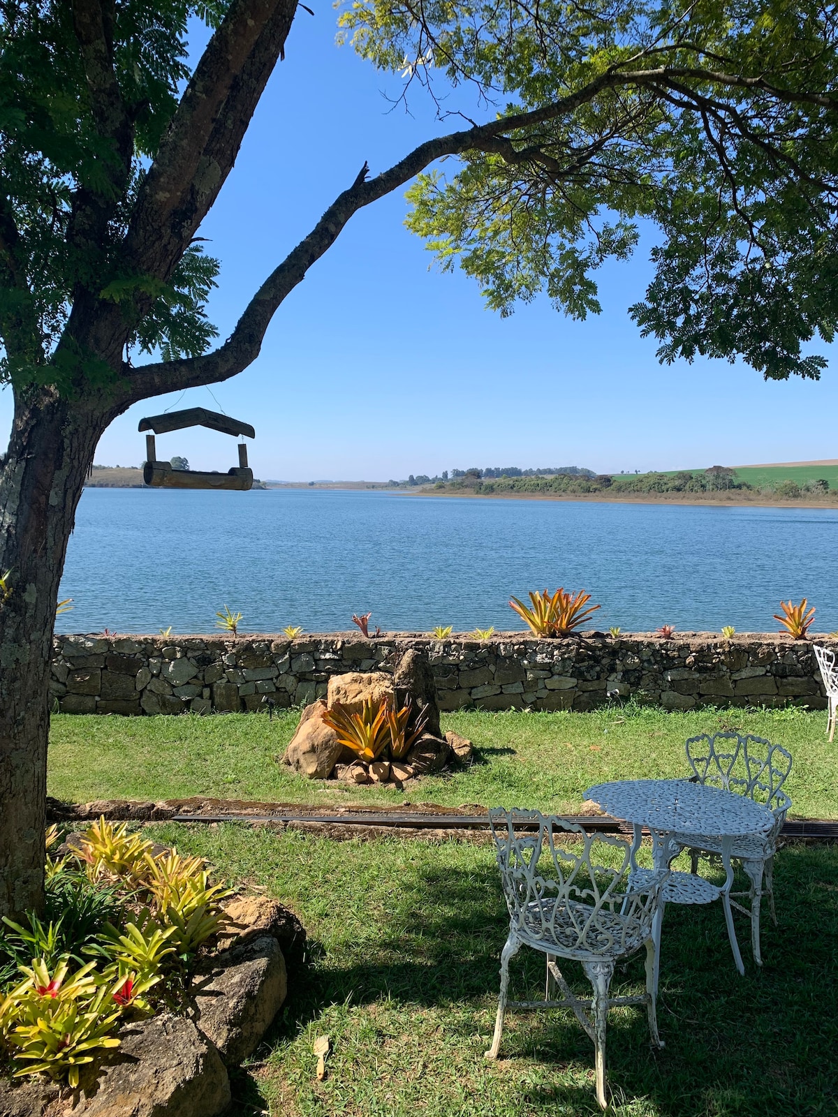 A serene lakeside view is framed by lush greenery, showcasing a stone wall and colorful plants. A small, white outdoor table and chairs are positioned under a tree, inviting relaxation and contemplation by the water.