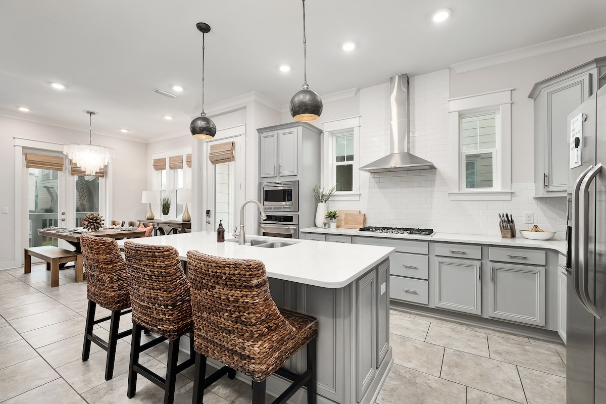 A modern kitchen is seen with a central island featuring a white countertop and seating for three on woven barstools. Stainless steel appliances are positioned against gray cabinetry, while large windows provide ample natural light, illuminating the tile flooring and contemporary design.