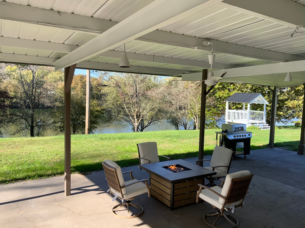 A covered patio area features a fire pit surrounded by four cushioned chairs, providing a comfortable gathering space. A grassy area extends to the river, with trees visible in the background. A grill stands nearby, and a gazebo is positioned by the water.