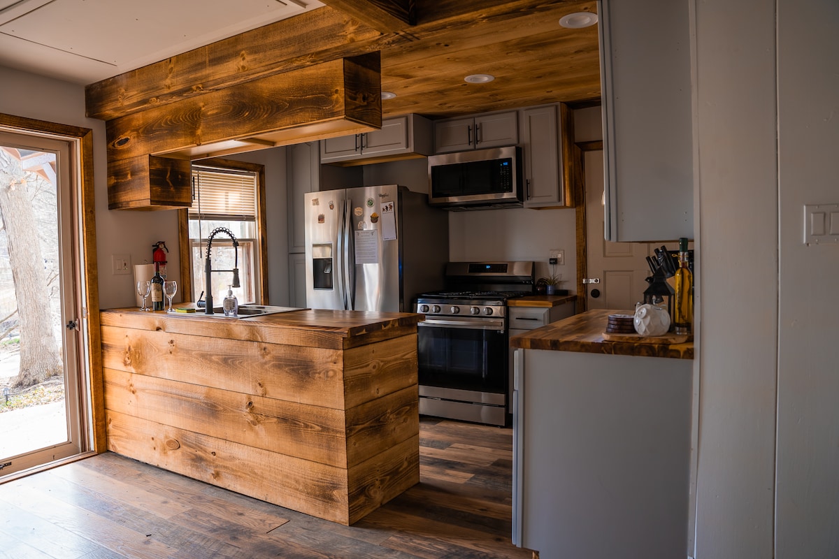 The kitchen features a wooden island with a quartz countertop, surrounded by stainless steel appliances. A large window allows natural light to enter, illuminating the modern cabinetry and sleek finishes. The wooden accents create a warm yet contemporary environment.