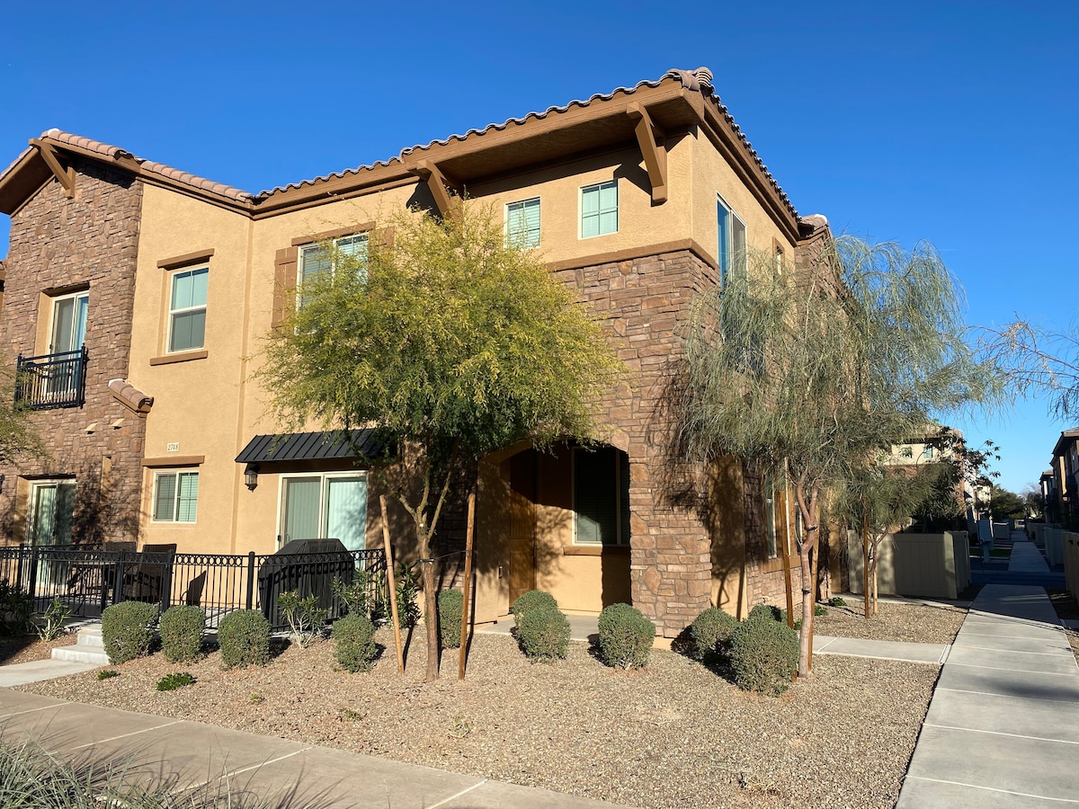 The townhouse exterior is characterized by a combination of stone and stucco finishes. Large windows are framed by greenery and ornamental shrubs, while the entrance is marked by a covered porch. Clear blue skies enhance the welcoming appearance of the corner unit.