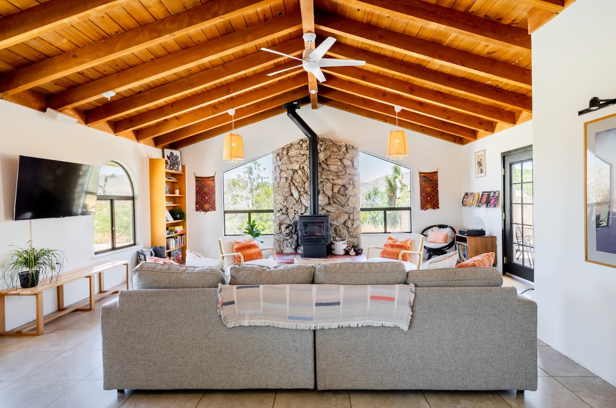 A spacious living area is characterized by a high wooden ceiling with exposed beams. Soft seating is arranged around a stone fireplace, with large windows framing views of the outdoors. Natural light fills the space, enhanced by warm light fixtures.