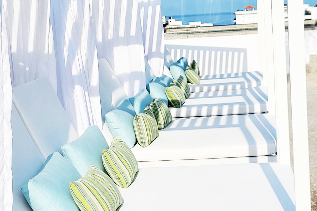 A row of sunbeds is positioned under draped white curtains, providing a relaxing space. Soft blue and striped green cushions are arranged neatly, creating a comfortable ambiance. The backdrop features a clear sky and distant buildings, adding to the serene atmosphere.