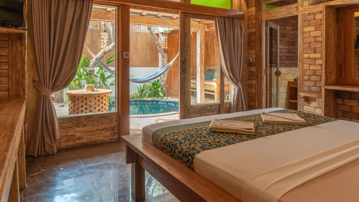 A spacious bedroom features a wooden bed with decorative pillows, offering views of a private patio with a hammock and a pool. Natural light fills the room through large glass doors, framed by elegant curtains that create a cozy atmosphere.