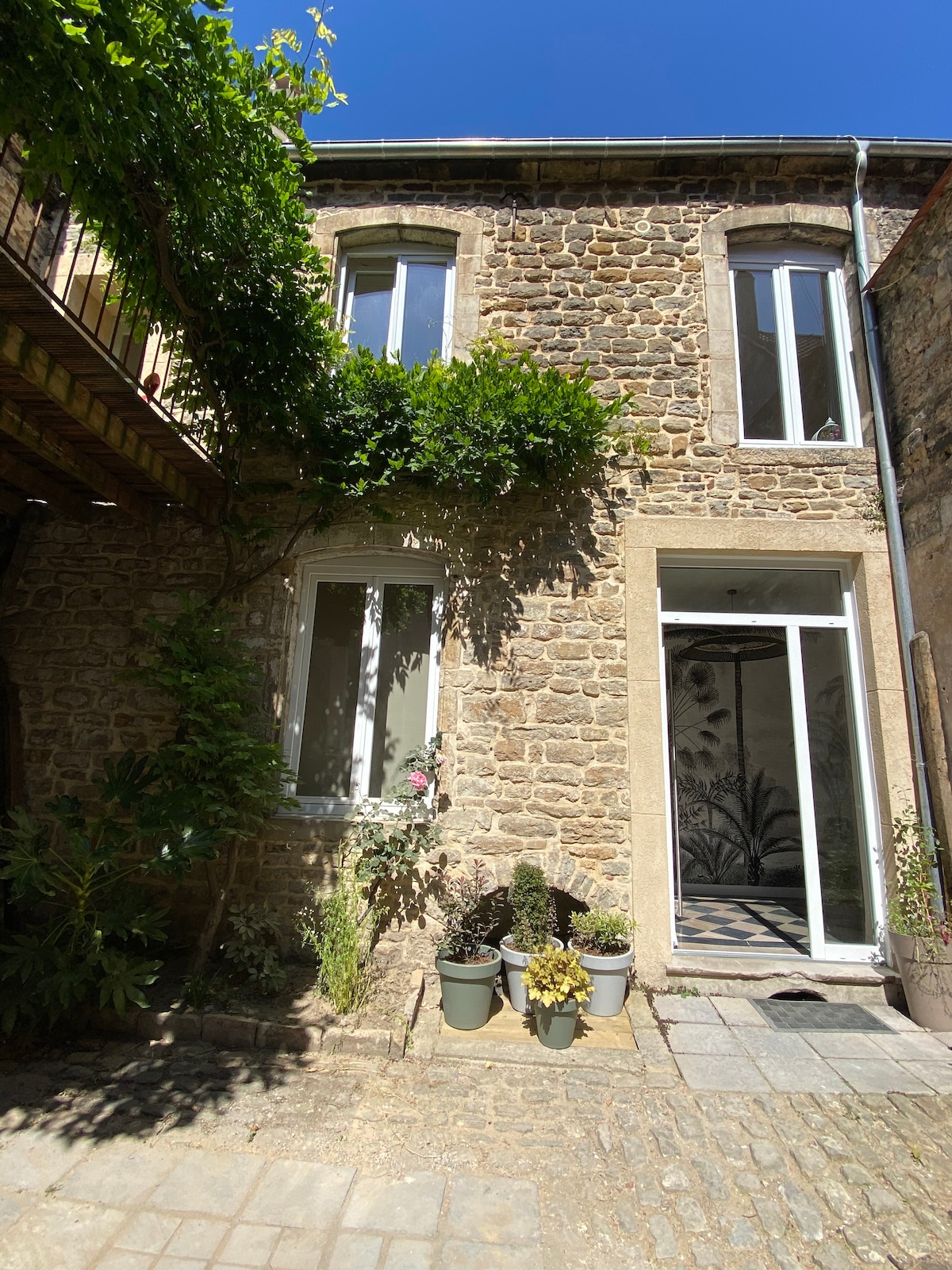 The exterior of a stone building features large windows framed by greenery. A spacious entrance with a glass door is visible, leading to a small stone pathway adorned with potted plants and flowers. Sunlight highlights the natural textures of the stone façade.