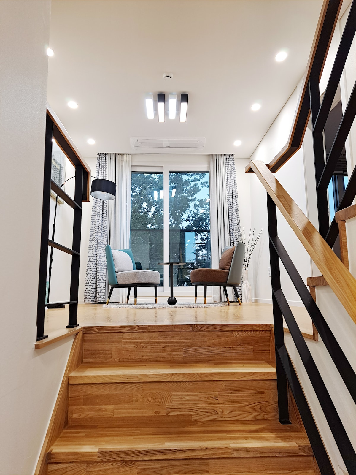 A staircase leads to a bright landing area with two chairs positioned beside a small table. Natural light streams through a large window adorned with curtains, illuminating the space. The modern decor features wooden accents and contemporary lighting.