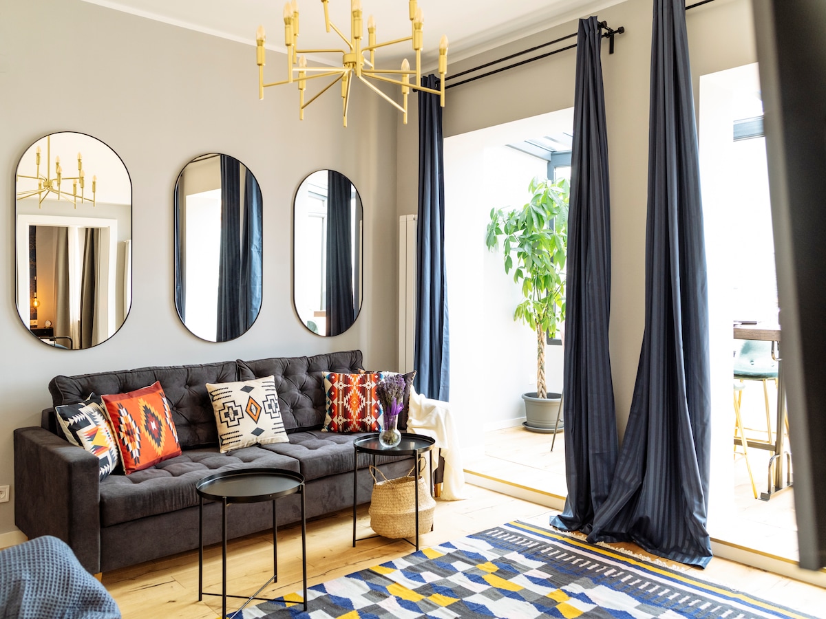 A comfortable living room is presented with a dark sectional sofa adorned with patterned cushions. Three round mirrors are mounted on the wall, reflecting light. A tall plant is visible near large windows, and a patterned area rug lays on the wooden floor.