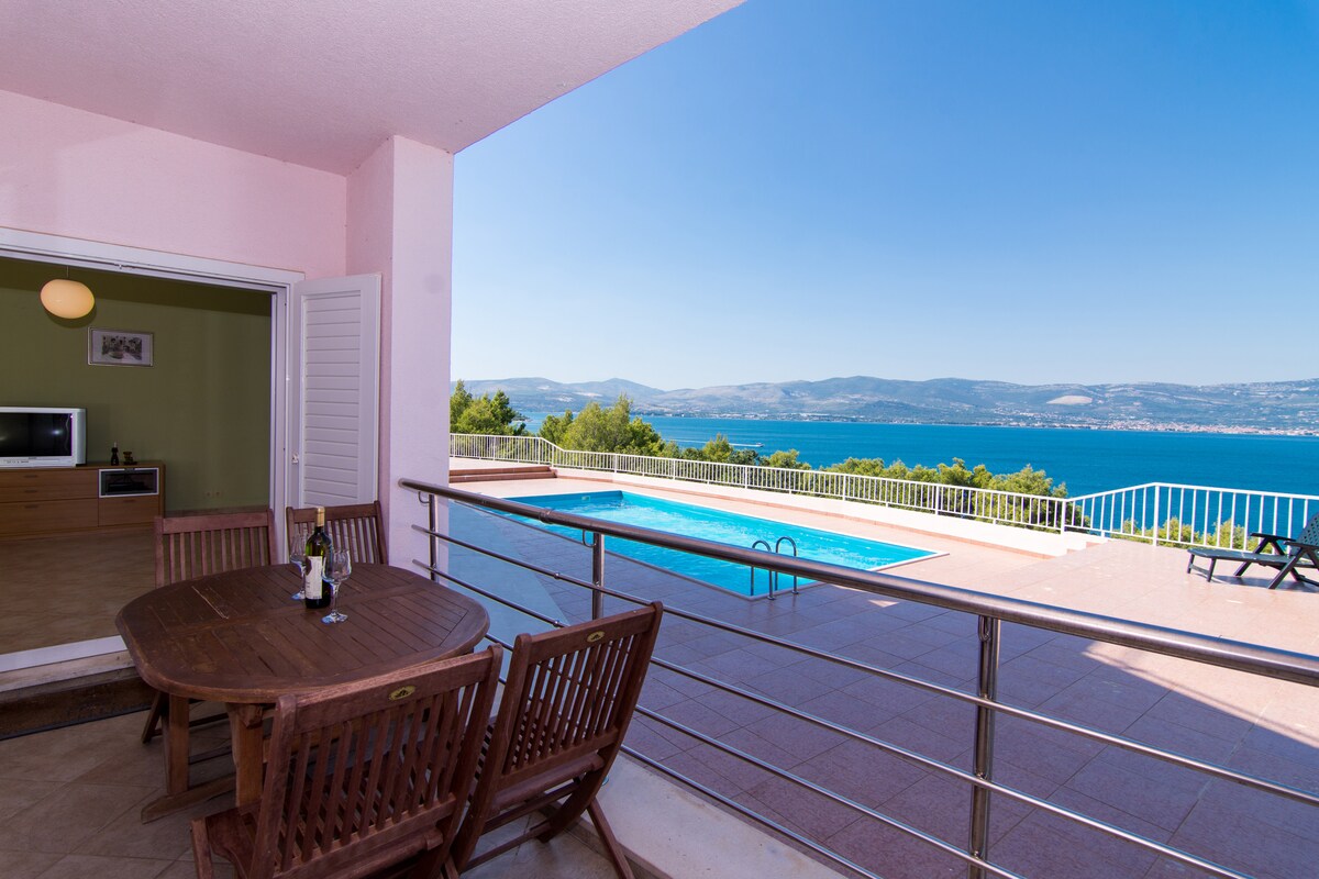 A terrace with a wooden dining table and chairs overlooks a swimming pool and the sea. Expansive views of the water and distant mountains are visible, with clear blue skies enhancing the scene. The interior is accessible through sliding glass doors.