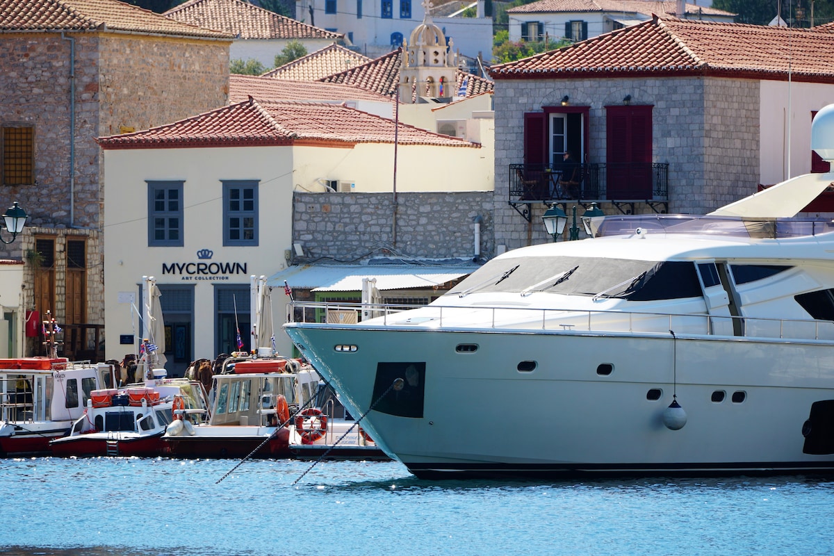 A view of the port features a sleek yacht moored alongside smaller boats. The MyCrown building, characterized by its traditional architecture, stands prominently in the background with its name displayed. Red rooftops and whitewashed walls create a charming coastal atmosphere.
