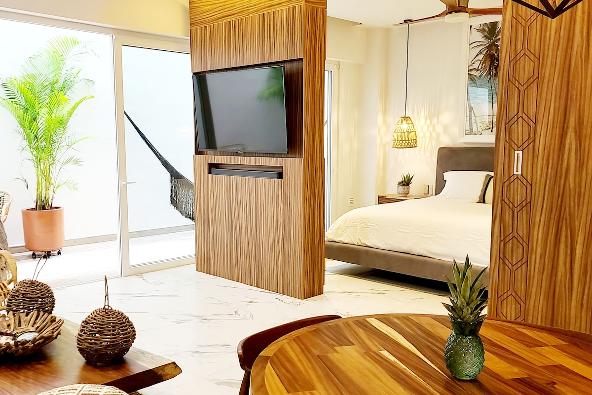 A modern living space with natural wood accents features a spacious bed behind a stylish wooden partition. A flat-screen TV is mounted on the wall, and light filters in through large sliding glass doors, which open to a private balcony adorned with a tropical hammock.