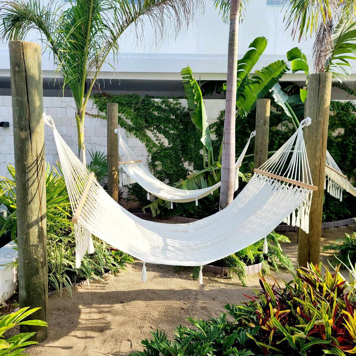 Two white hammocks are suspended between wooden posts in a lush garden setting. Tropical plants, including palms and leafy greenery, surround the area, creating a serene atmosphere on a sandy surface.