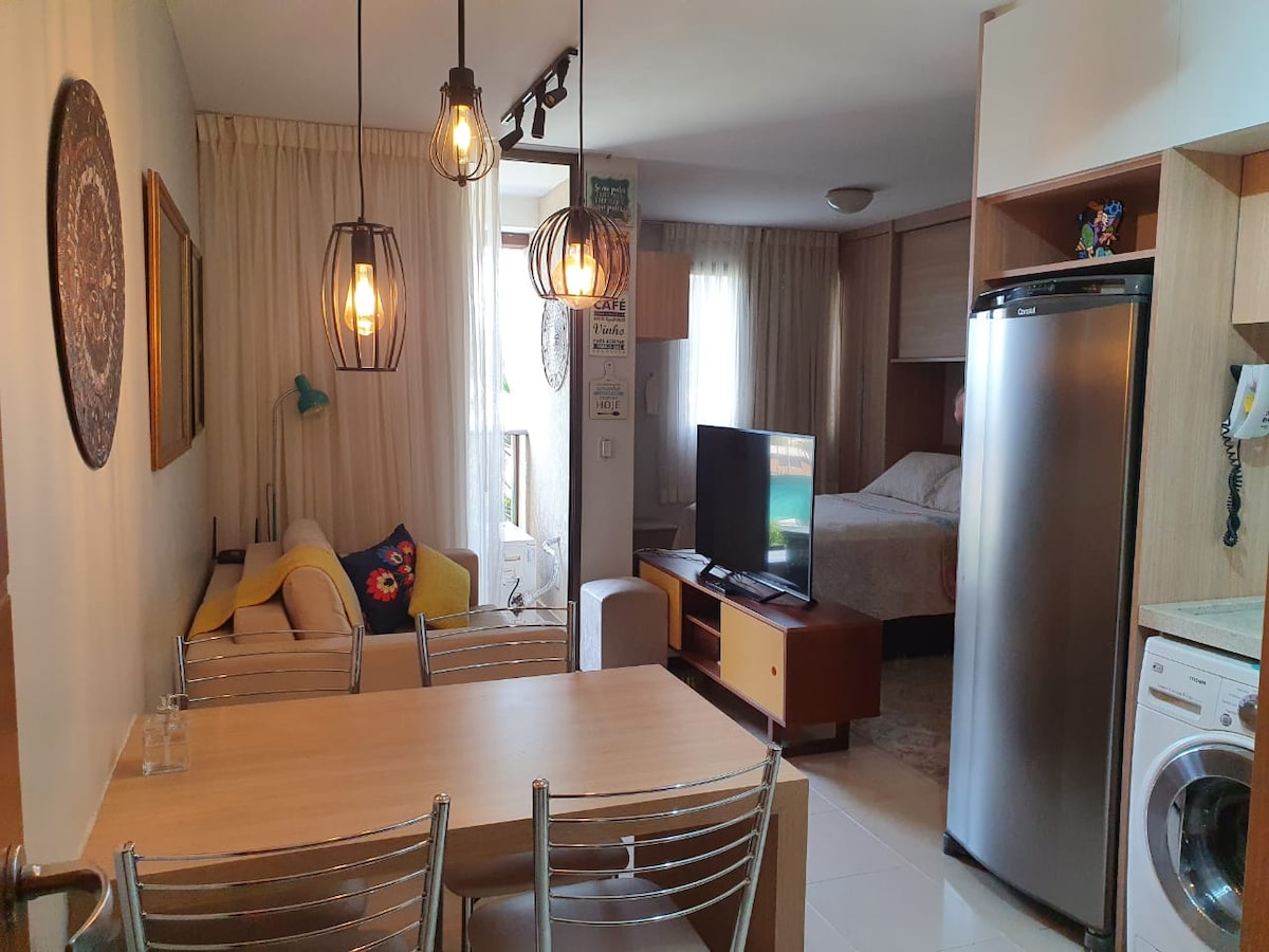 An inviting living area features a modern dining table with metallic chairs, illuminated by stylish pendant lights. A cozy seating arrangement is visible, along with a flat-screen TV mounted on a wooden unit. A spacious bed is situated in the background, creating a functional space.