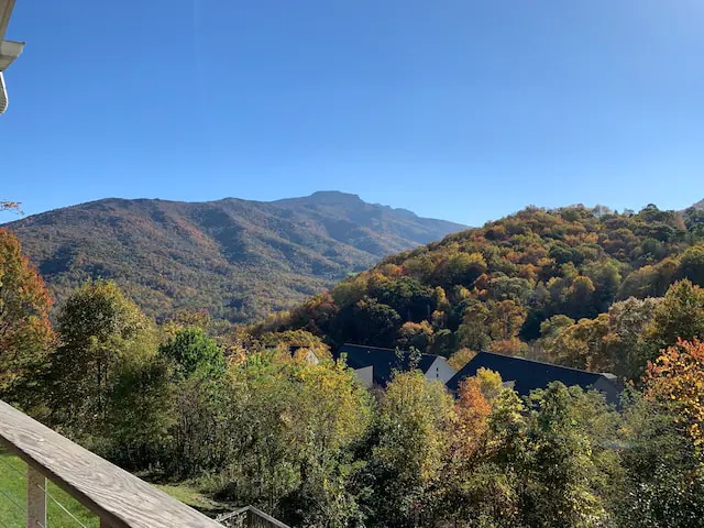 A stunning mountain view showcases colorful autumn foliage, with rolling hills and peaks in the background under a clear blue sky. Green trees and patches of orange and gold frame the landscape, creating a serene and natural ambiance.