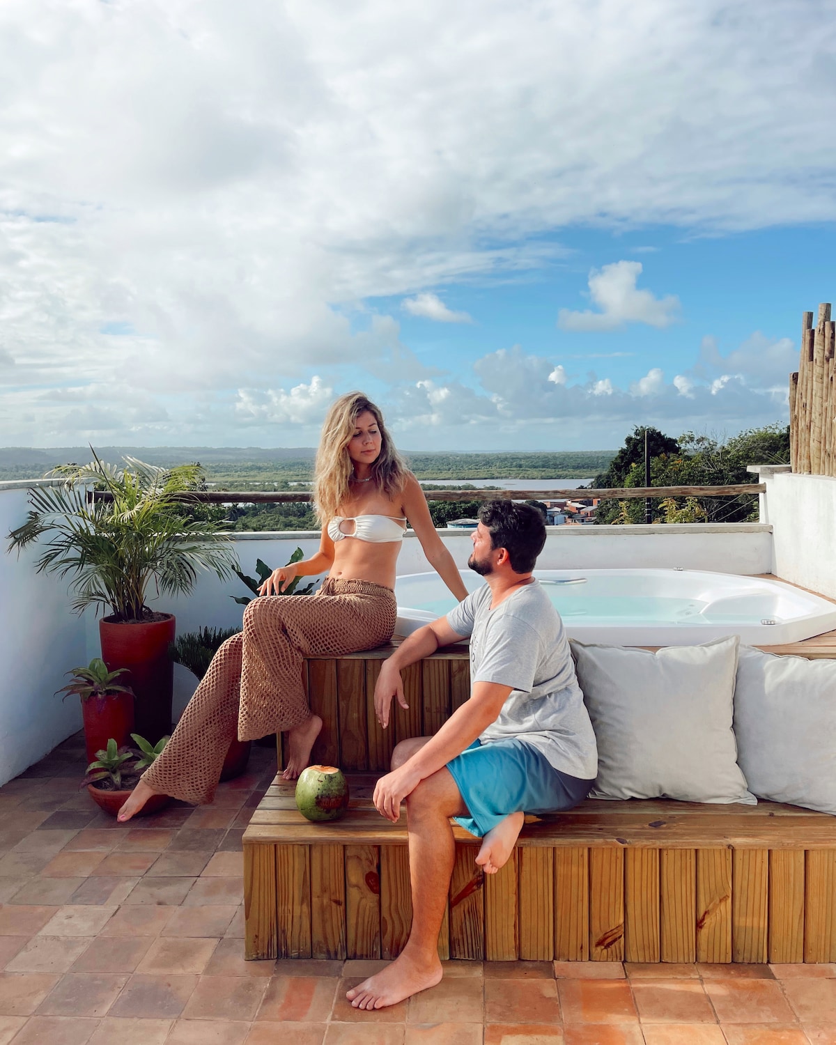 An inviting terrace features a private jacuzzi overlooking a scenic view, surrounded by plants. Comfortable seating in natural wood invites relaxation. A partly cloudy sky adds to the tranquil atmosphere, creating a peaceful outdoor space.