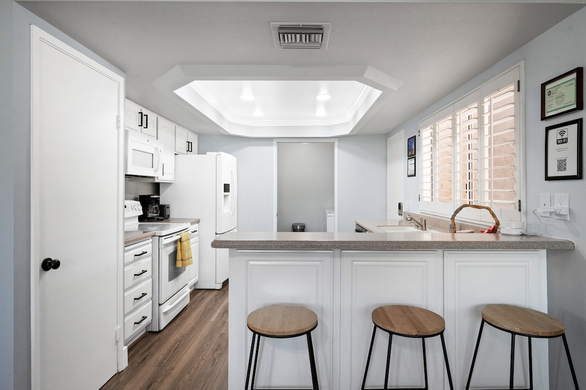 A well-equipped kitchen features white cabinetry and modern appliances, including a refrigerator and stove. A central island with three wooden stools provides seating. Natural light enters through the large windows adorned with shutters, creating a bright and functional workspace.