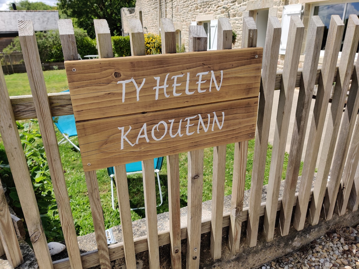 A wooden sign is displayed prominently, featuring the text 'TY HELEN KAOUENN' in a simple, elegant font. The sign is mounted on a wooden fence, surrounded by green grass and some garden elements, contributing to a welcoming entrance to the property.