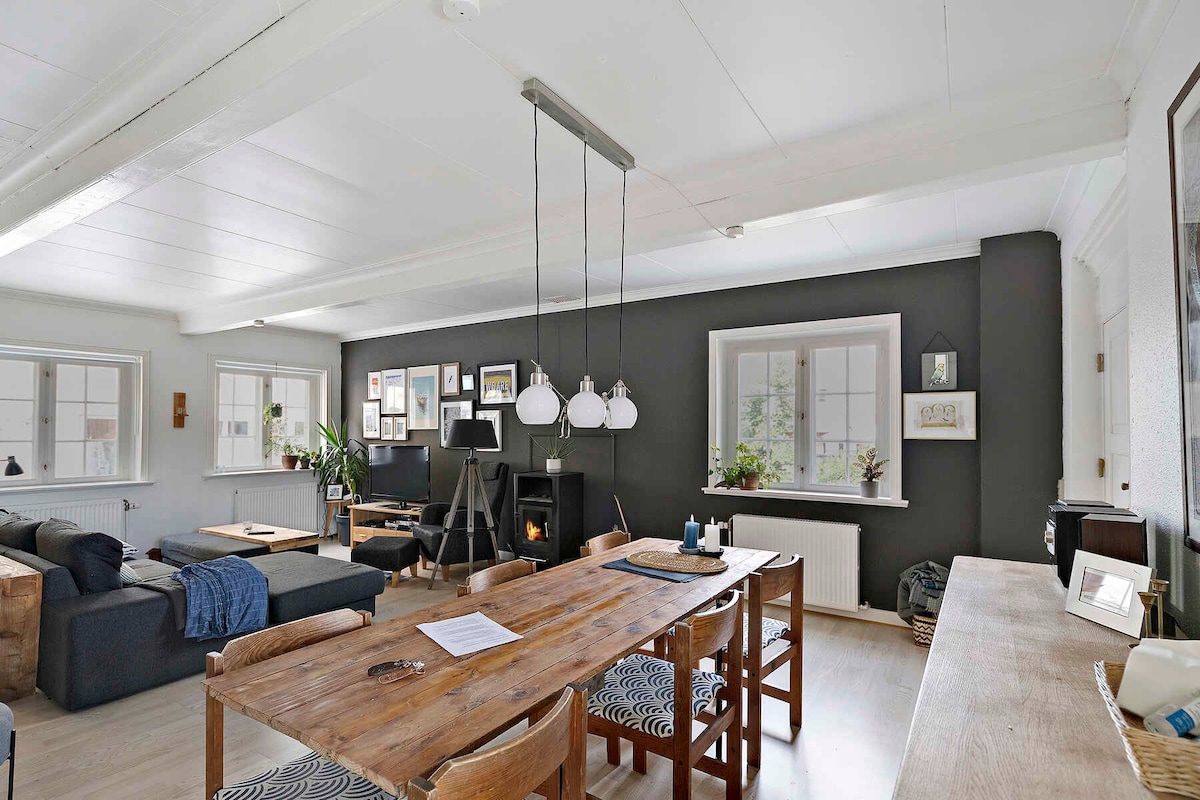 The living area features a rustic wooden dining table surrounded by patterned chairs, set against a backdrop of dark walls adorned with framed artwork. A comfortable seating arrangement is visible, along with natural light streaming in through the large windows, enhancing the room's cozy ambiance.