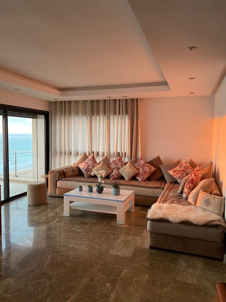 A spacious living area features a large sectional sofa adorned with decorative cushions, complemented by a coffee table. Large glass doors provide an unobstructed view of the ocean, with natural light softly illuminating the room. A neutral color palette enhances the serene ambiance.